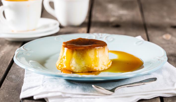 Fotografia em tons de branco e amarelo de uma bancada cinza, com um pano branco dobrado, uma colher de inox de sobremesa, um prato de sobremesa azul e nele um mini pudim redondo com calda de caramelo. Ao fundo, duas xícaras brancas com pudim dentro.