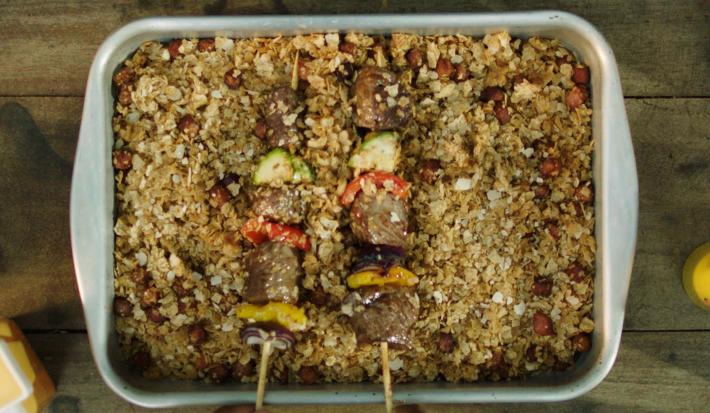 Fotografia em tons de marrom em uma bancada de madeira, um recipiente branco retangular com a farofa de Neston e bacon, por cima, espetinhos de carne e legumes para acompanhar a farofa.