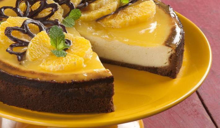fotografia em tons de roxo, branco e amarelo de uma bancada roxa vista de cima. Contém um suporte para servir sobremesa cor amarelo com uma torta e fatias de laranja por cima.