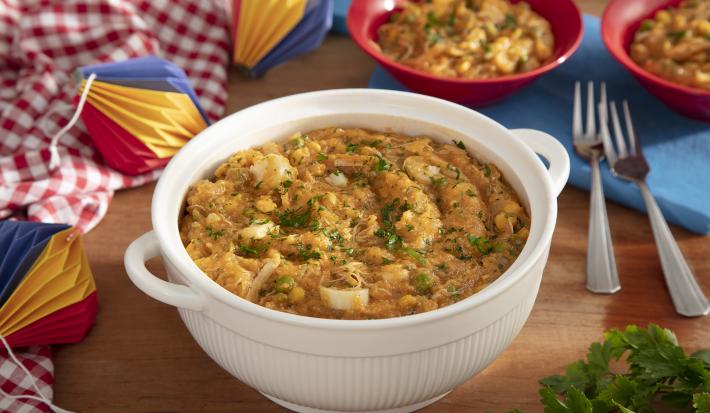 Foto da receita de Mungunzá de festa junina. Observa-se uma tigela grande branca com a receita dentro e, atrás, dois pratos fundos vermelhos cheios. Decoração de festa junina com tecidos e balões de papel decoram a foto do lado esquerdo