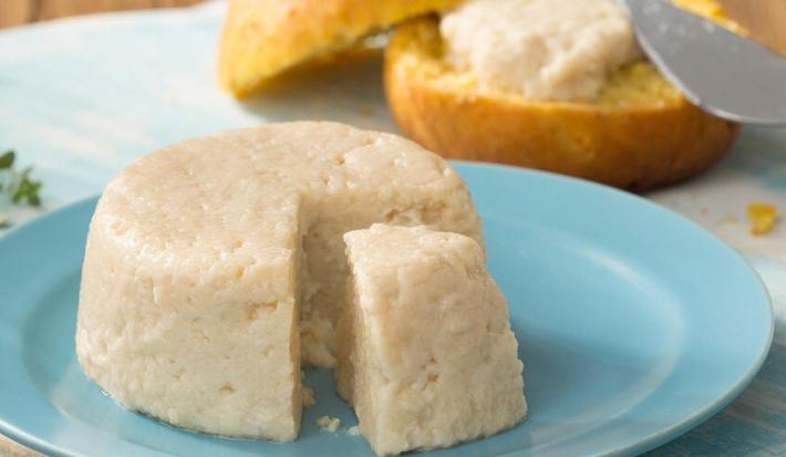 Fotografia em tons de azul e branco de uma bancada azul claro vista de frente, um prato redondo e azul contém um queijo redondo com uma fatia retirada. Ao fundo um pão redondo cortado ao meio e uma espátula com creme colocando em uma das fatias do pão.