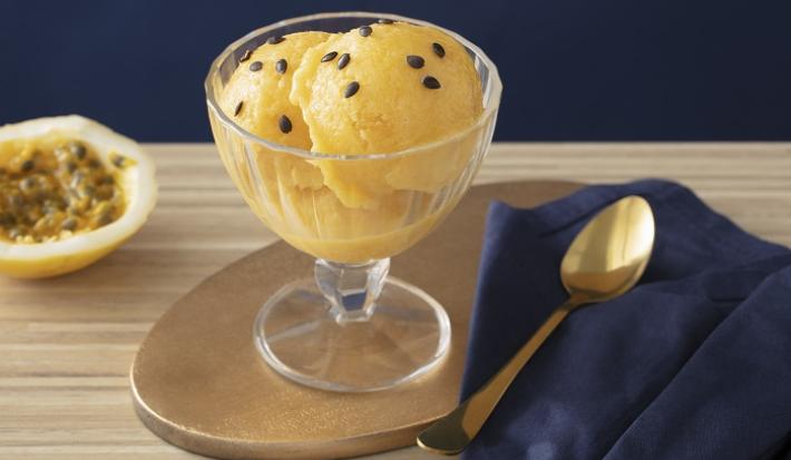 Foto em tons de amarelo da receita de sorbet de damasco e maracujá servida em uma taça de vidro sobre uma mesa de madeira com uma colher prateada ao lado sobre um pano de veludo azul. Ao fundo ainda há metade de um maracujá