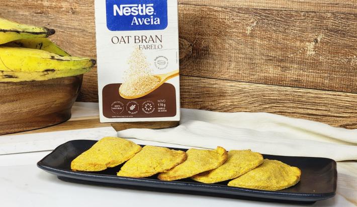 Fotografia em tons de marrom em uma bancada de madeira marrom, uma régua preta ao centro com pastéis de banana recheados com chocolate dentro. Ao fundo, embalagem da Nestlé Farelo de Aveia e um cacho de banana.