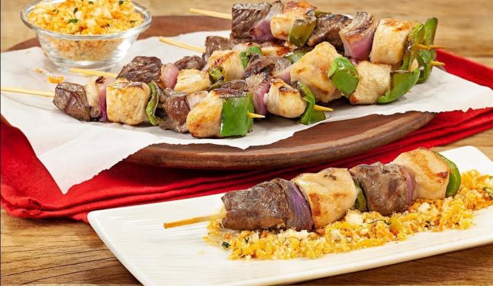 Fotografia em tons de marrom em uma bancada de madeira com um pano vermelho, um prato marrom com espetinhos de carne e frango em cima dele. Ao fundo, um potinho de vidro com farofa.