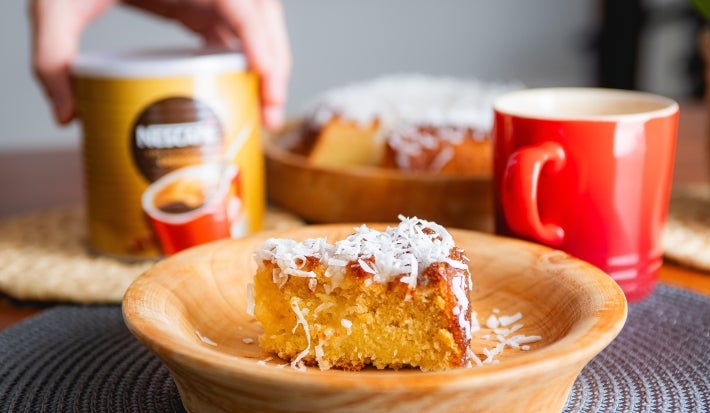 Fotografia mostra uma fatia de bolo com coco ralado em cima. Ao fundo uma lata de nescafé e uma xícara vermelha