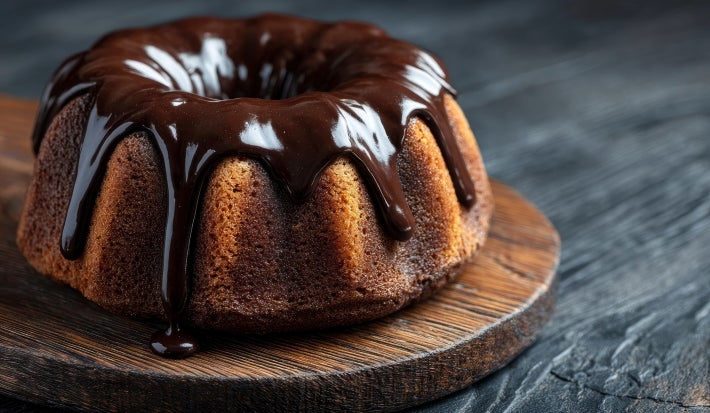 Fotografia de um bolo bundt com crosta dourada, coberto por um glaze de chocolate escuro, posicionado em uma tábua de madeira rústica.
