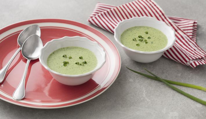 Fotografia em tons de vermelho em uma bancada de madeira cinza com um pano com listras vermelhas, um potinho branco pequeno com a sopa de alface. Ao lado, um prato vermelho grande com um potinho branco em cima dele e a sopa de alface dentro.