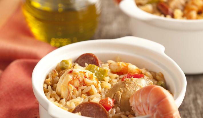 fotografia em tons de cinza e branco de uma bancada vista de frente, contém dois potinhos brancos e redondos com arroz, pedaços de carne suína e frutos do mar. Ao lado um pano laranja com dois garfos para servir e uma jarra de azeite