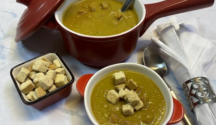 \foto em tons de verde da receita de creme de ervilha servida em uma panelinha marrom com uma colher de servir prateada dentro, à frente há um bowl laranja e branco com uma porção do creme e ao lado há crouttons e uma colher prateada