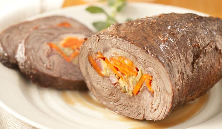 fotografia em tons de branco e laranja de uma bancada vista de frente. Contém um pano branco, um prato redondo com uma picanha recheada com cenoura, azeitona e bacon. Ao fundo um pote com um molho.