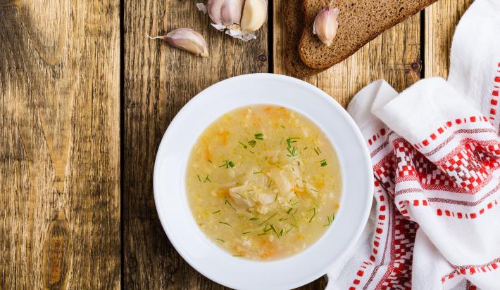 Fotografia vista de cima de uma sopa de mandioquinha, cenoura e vagem em um prato redondo na cor branca. Ao lado, uma toalha de mesa branca com detalhes em vermelho, e alguns dentes de alho ao lado de duas fatias de pão sobre uma mesa de madeira.