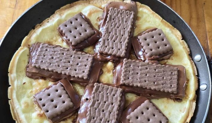 Fotografia em tons de marrom e vermelho com um prato preto ao centro. Em cima do prato tem uma pizza feita de massa de ovo e tapioca. Por cima da massa existe vários biscoitos Choco Trio derretidos.