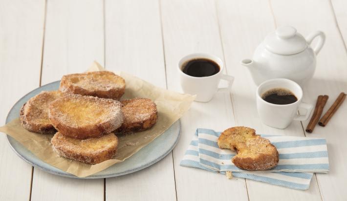Fotografia em tons de dourado e branco, com rabanadas fritas sobre papel manteiga e prato azul, ao lado serviço de café, paus de canela e rabanada sobre guardanapo listrado em azul e branco, tudo sobre bancada em tom pastel.