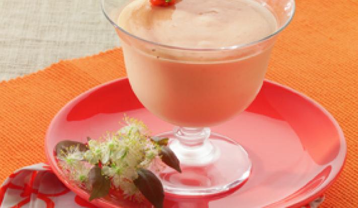 Fotografia em tons de laranja em uma mesa de madeira com uma toalha laranja, um pano vermelho desenhado com flores, um prato vermelho e uma taça de vidro com a mousse de pitanga dentro e uma pitanga em cima para decorar.