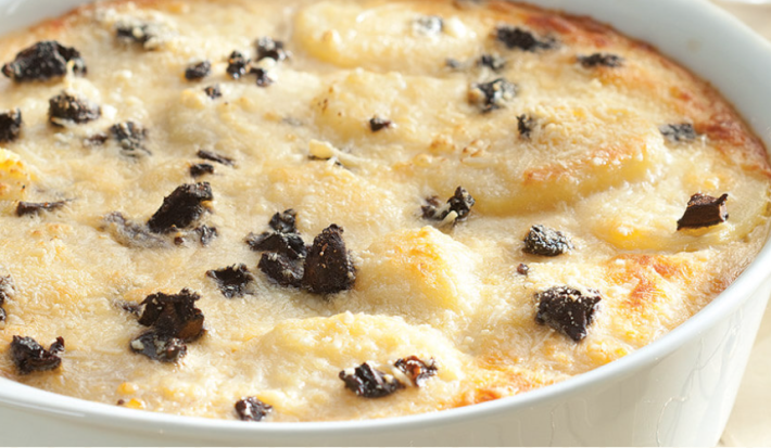 Fotografia em tons pastel, branco e dourado, ao centro refratário com batatas gratinadas com funghi sobre toalha com decoração dourada, ao fundo copos dourados e pilha de pratos brancos com guardanapo vermelho em cima.