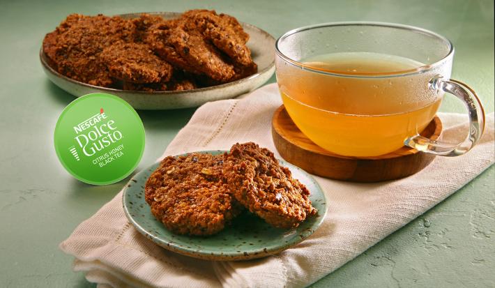 Fotografia em tons de verde em uma bancada de cimento queimado, um pano rosinha com um prato pequeno e os biscoitos de ameixa com aveia. Ao lado, uma xícara de vidro com o chá cítrico e a cápsula verde da Dolce Gusto do Chá Cítrico.