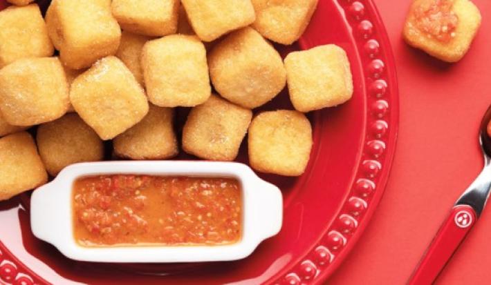 Fotografia em tons de vermelho em uma mesa vermelha, com um prato vermelho raso grande e vários dadinhos de polenta recheados com queijo coalho. Ao lado, um potinho com o molho de pimenta biquinho.