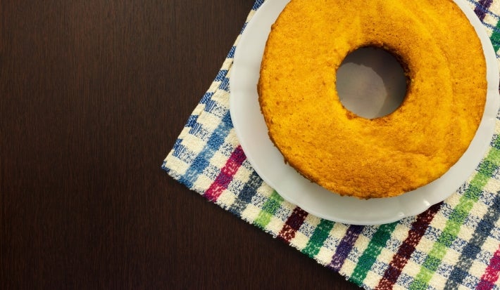 Um bolo de cenoura inteiro, redondo e simples, está colocado sobre um prato branco. A base do prato está sobre um pano de prato colorido xadrez, e o fundo é uma mesa escura de madeira, criando um contraste bonito com o bolo dourado.