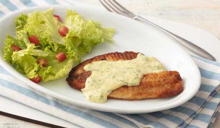Fotografia em tons de azul em uma bancada de madeira clara com um pano listrado azul claro e um prato oval branco com o filé de pescada com molho de limão e mostrada e salada de alface com tomates.