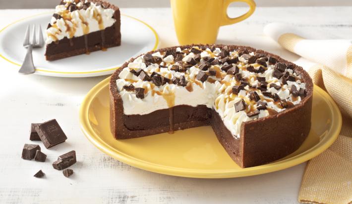 Fotografia em tons de amarelo em uma bancada branca com um prato grande amarelo e a torta de chocolate com caramelo em cima cortada ao meio. Ao lado, um prato branco com uma fatia da torta de chocolate, uma xícara amarela e pedaços de chocolates.