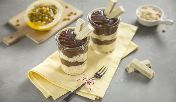 Fotografia em tons de amarelo em uma bancada cinza com um pano amarelo ao centro com dois potinhos de vidro com o bolo de pote de brigadeiro com dois KitKat de Passion Fruit decorando o pote. Ao fundo, um maracujá cortado ao meio.