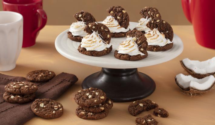 Foto de uma bancada com canecas vermelha e brancas, um tecido marrom, pedaços de coco seco. Ao centro há um prato com as receitas de cookies com merengue de coco e mais alguns Cookies Prestígio espalhados na bancada