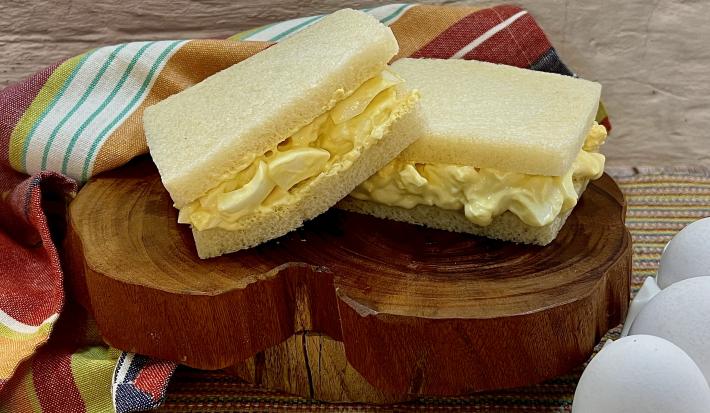 Foto da receita de Sanduíche de Ovos. Observa-se uma tábua de madeira com dois sanduíches de pão de forma sem casca com o recheio de ovos dentro sobre a tábua.