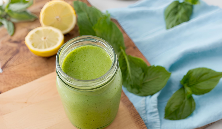 Fotografia de um copo de vidro decorativo com suco detox de cor verde feito com couve, maçã verde, pepino, gengibre e nutren. O copo está sobre uma tábua de madeira, ao lado de folhas de hortelã e metades de limão siciliano sobre a mesa.