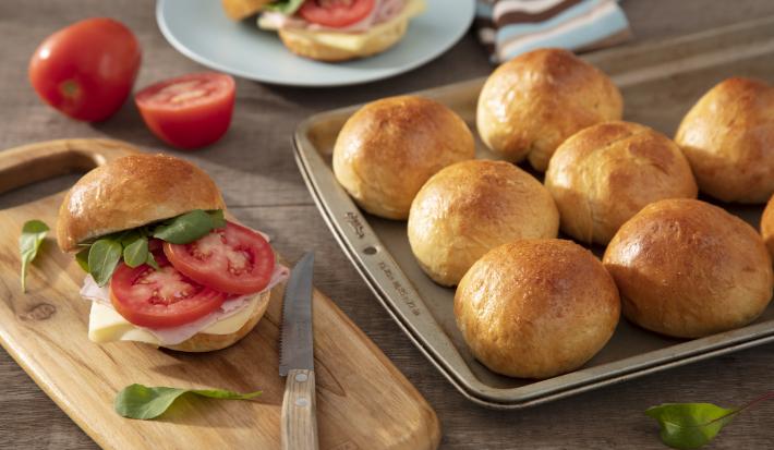 Fotografia em tons de marrom em uma bancada de madeira escura, uma tábua de madeira com um sanduíche de pão de batata doce recheado com presunto, queijo e rodela de tomate. Ao lado, uma forma com vários pãezinhos.