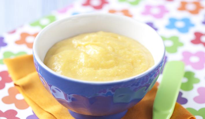 Fotografia em tons de azul em uma bancada de madeira azul clara, um pano com flores coloridas, um paninho laranja, um recipiente azul com a papinha dentro dele. Ao lado, uma colher infantil verde.