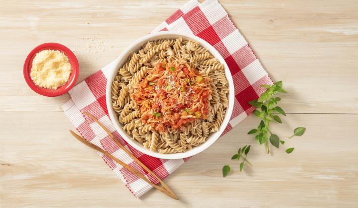 Fotografia em tons de vermelho em uma bancada de madeira com um pano de xadrez vermelho, em cima um prato fundo redondo com o macarrão integral com molho de frango dentro. Ao lado, um pegador de macarrão e um potinho vermelho com queijo ralado.
