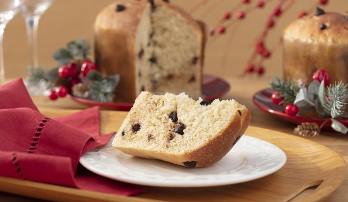 Imagem de uma fatia de panetone caseiro com gotas de chocolate em um prato branco. Ao fundo, o panetone e decorações de Natal.