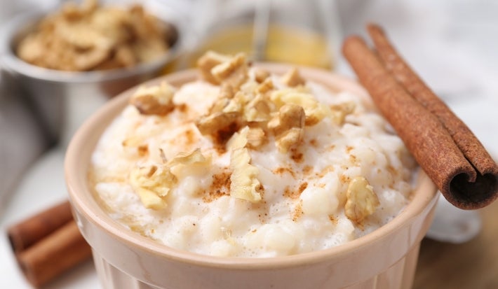 Em cima de uma mesa, recipiente redondo com arroz doce cremoso, com uma textura suave e aveludada, decorado com nozes picadas e canela em pau.