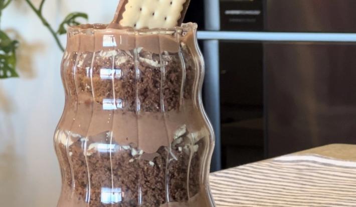 Fotografia de um recipiente decorativo grande com um pavê com camadas de bolo e creme de whey, também com chocotrio de amendoim. Por cima tem uma unidade de choco trio de amendoim.