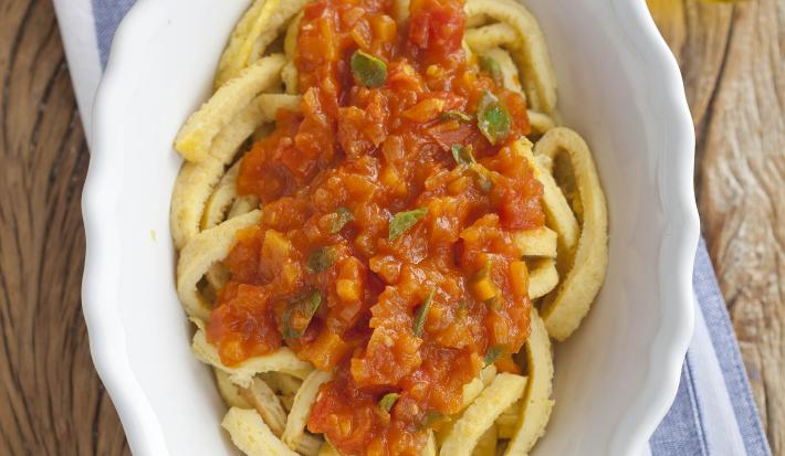 fotografia em tons de marrom, branco, bege e vermelho de uma bancada marrom vista de cima, ao centro um pano listrado em tons de azul e branco com um recipiente branco oval com macarrão e molho de tomate por cima