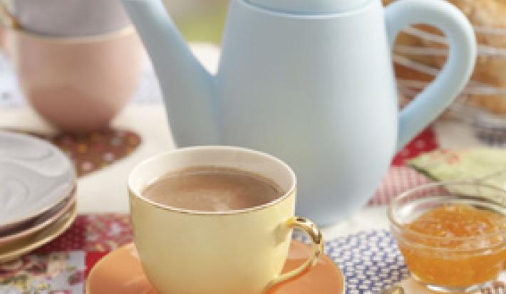 foto em tons de marrom, bege e azul tirada de um pratinho bege com uma xicara bege que contém o chocolate quente dentro. Ao fundo um bule azul