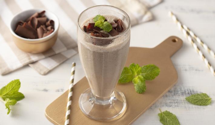 Fotografia em tons de marrom e branco de uma bancada, com uma tábua de madeira e um copo sobre ela com a bebida de chocolate decorada com raspas de chocolate e folha de hortelã. Ao redor potinho com raspas de chocolate e folhas de hortelã.
