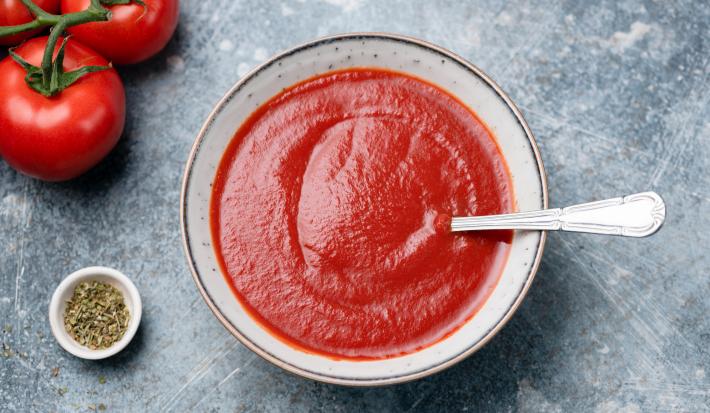 fotografia em tons de cinza e vermelho de uma bancada cinza vista de cima. Ao centro um recipiente redondo com sopa de tomate e um colher para servir, e ao lado esquerdo contém tomates.