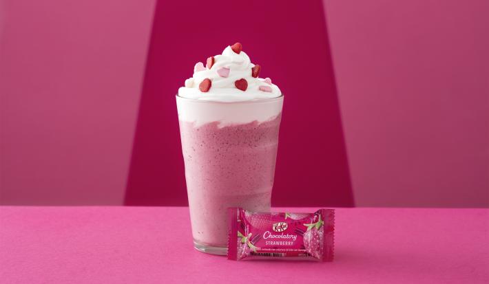 Foto da receita de Frappé. Observa-se um fundo rosa com um copo alto decorado de chantily e confeitos de coração com um kit kat de morango na frente.