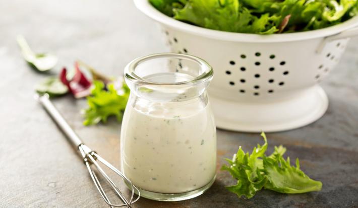Imagem de um pode de vidro com molho de iogurte, um mini fuet e umas folhas de salada. Ao fundo, um escorredor branco com folhas de alface.