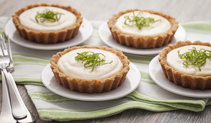 Fotografia em tons de verde em uma mesa de madeira com um pano listrado em verde e quatro pratinhos brancos cada um com a torta creme de limão. Ao lado, três garfos.