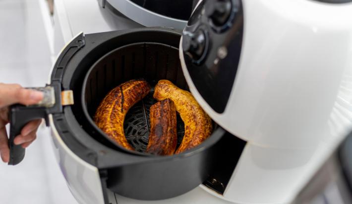 Fotografia dem tons claros de uma mão puxando a cestinha da air fryer branca com uma receita de banana douradinha.