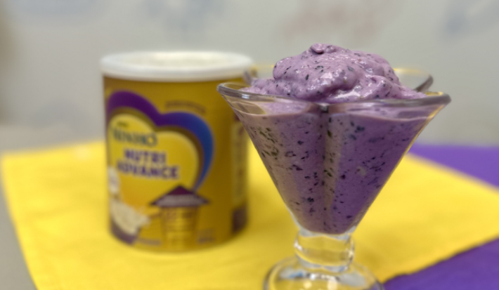 sobre um fundo azul e amarelo e branco está uma taça com um sorvete cremoso roxo cheio de pedacinhos de uva, ao fundo desfocado está uma lata do produto Ninho NutriAdvance em tons de dourado.