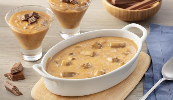 Fotografia em tons de bege em uma bancada de madeira com um pano azul ao lado, uma tábua de madeira, um recipiente oval branco com alças laterais e o strogonoff de KitKat dentro. Ao fundo, duas taças com o strogonoff e um pote com barrinhas de KitKat.