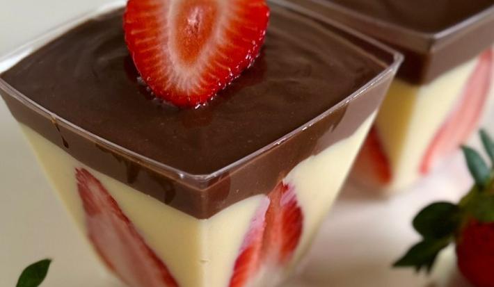 Fotografia de dois potes quadrados transparentes com bombom de morango com ganache de chocolate, morangos decorando a parede do copinho junto ao creme branco, e tem uma fatia por cima. Os potinhos estão sobre uma mesa branca, com morangos ao redor.