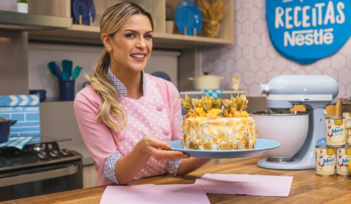 Foto da confeiteira Beca Milano segurando a receita de Bolo de Frutas Amarelas com Merengue, atrás de uma bancada de madeira de cozinha.