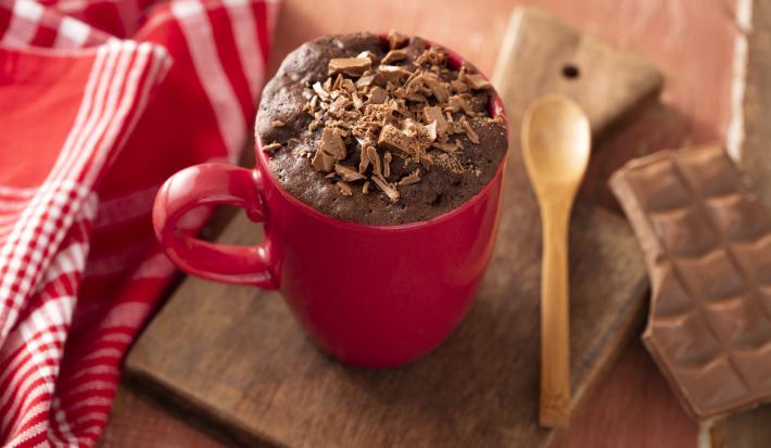 Fotografia em tons de vermelho de uma bancada de madeira, ao centro uma tábua de madeira com uma caneca vermelha e um bolo de chocolate com raspas de chocolate dentro. Ao lado uma pano vermelho com listras brancas e uma colher de madeira.