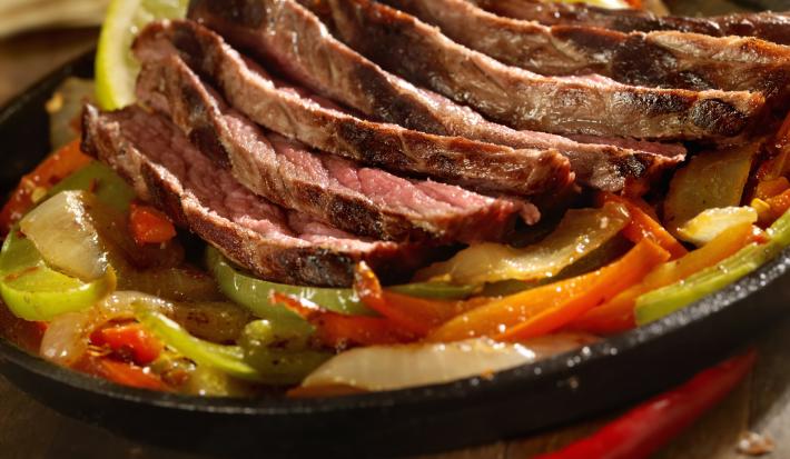 Fotografia de tirinhas de carne com cebola e pimentão temperadas. As carnes estão por cima dos legumes, que estão dentro de uma panela de ferro.
