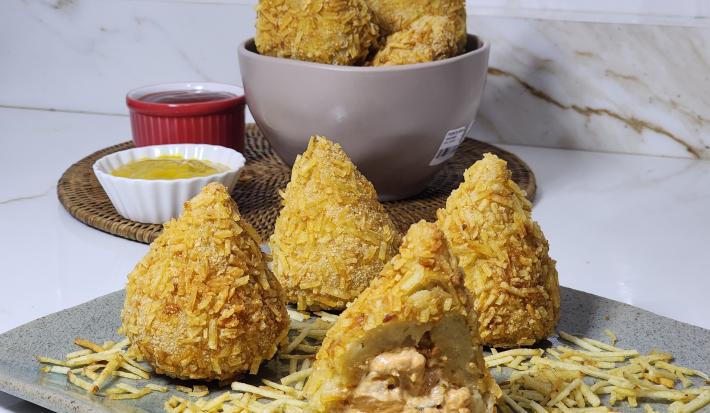 Foto da receita de coxinha de estrogonofe empanada em batata palha, servidas em um prato cinza, com potes ao fundo contendo ketchup e mostarda.