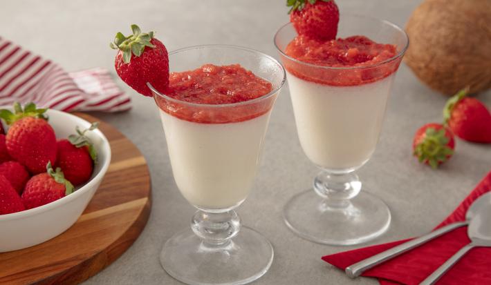 Foto da Receita de Taças de coco com purê de morango. Observa-se duas taças de vidro com o mousse e o purê com morangos decorando a borda da taça. Como decoração, vemos morangos ao lado direito em um potinho e um coco fresco ao lado direito